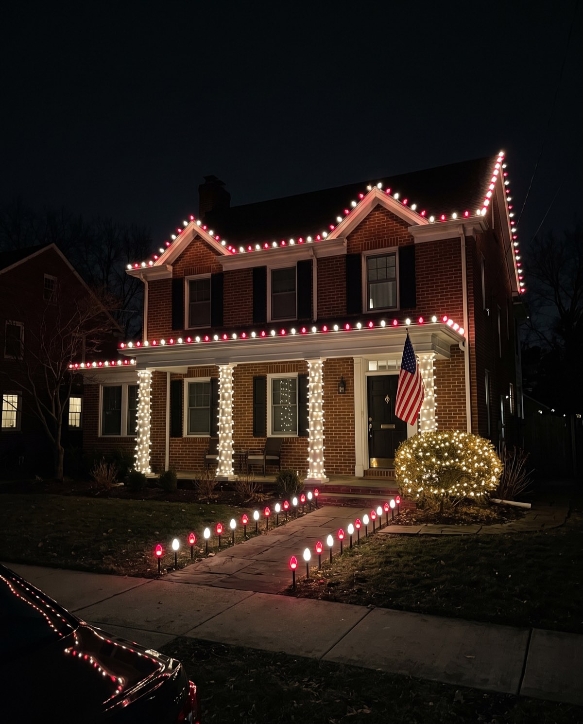 A brick two-story with alternating red and white C9 bulbs on the rooflines and red and white stakes lining the front walk