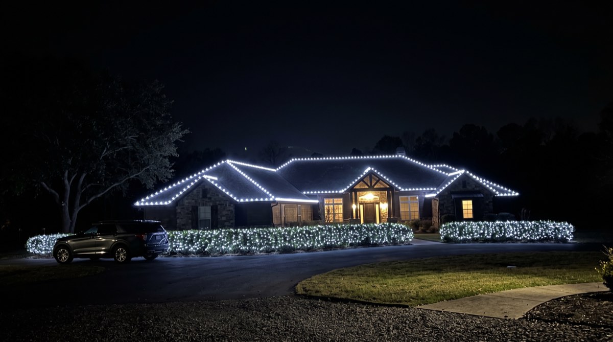 A stone ranch at night outlined in cool bright white C9 bulbs with a long lit hedge across the front