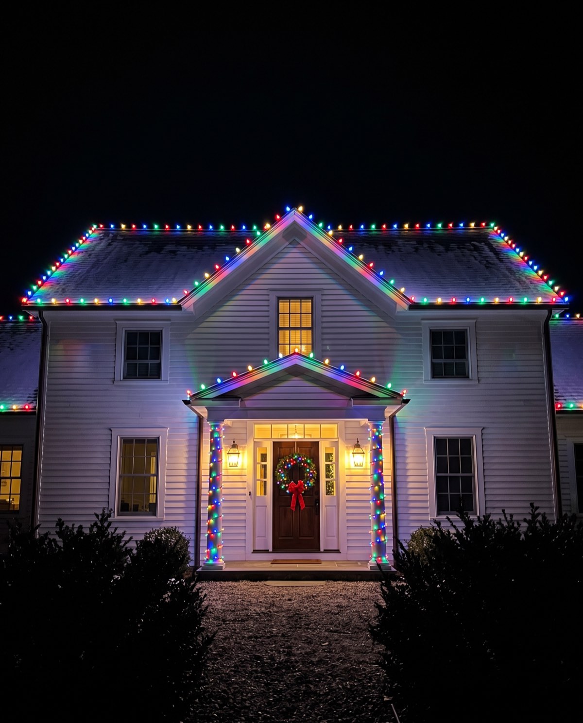 A white farmhouse colonial with classic multicolor C9 bulbs on the rooflines and multicolor wraps on the portico columns