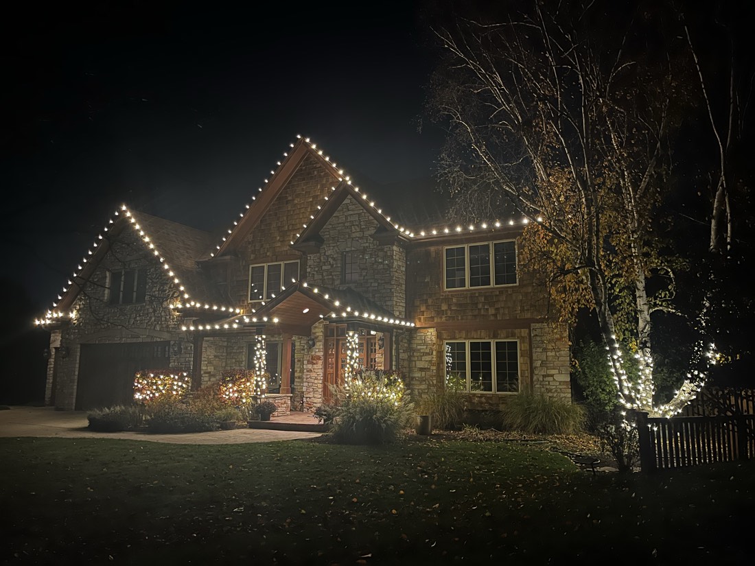 Stone and cedar-shake home with warm white C9 bulbs across every gable, shrubs and a birch tree wrapped in mini lights