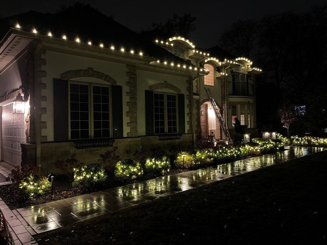 Stone French-country home on a rain-wet evening, rooflines and window arches in warm C9 bulbs with boxwood hedges lit along the walkway