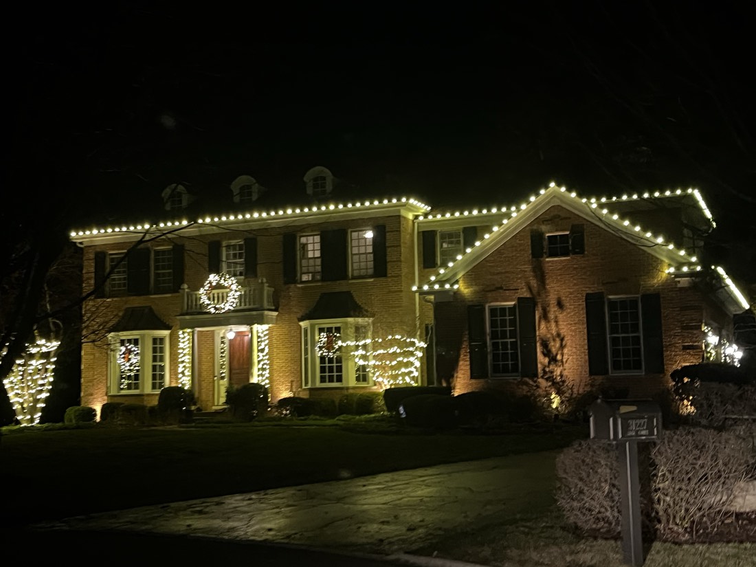 Brick Georgian colonial with dormers, rooflines in warm white C9 bulbs, garlands on the bay windows and a lit wreath over the entry