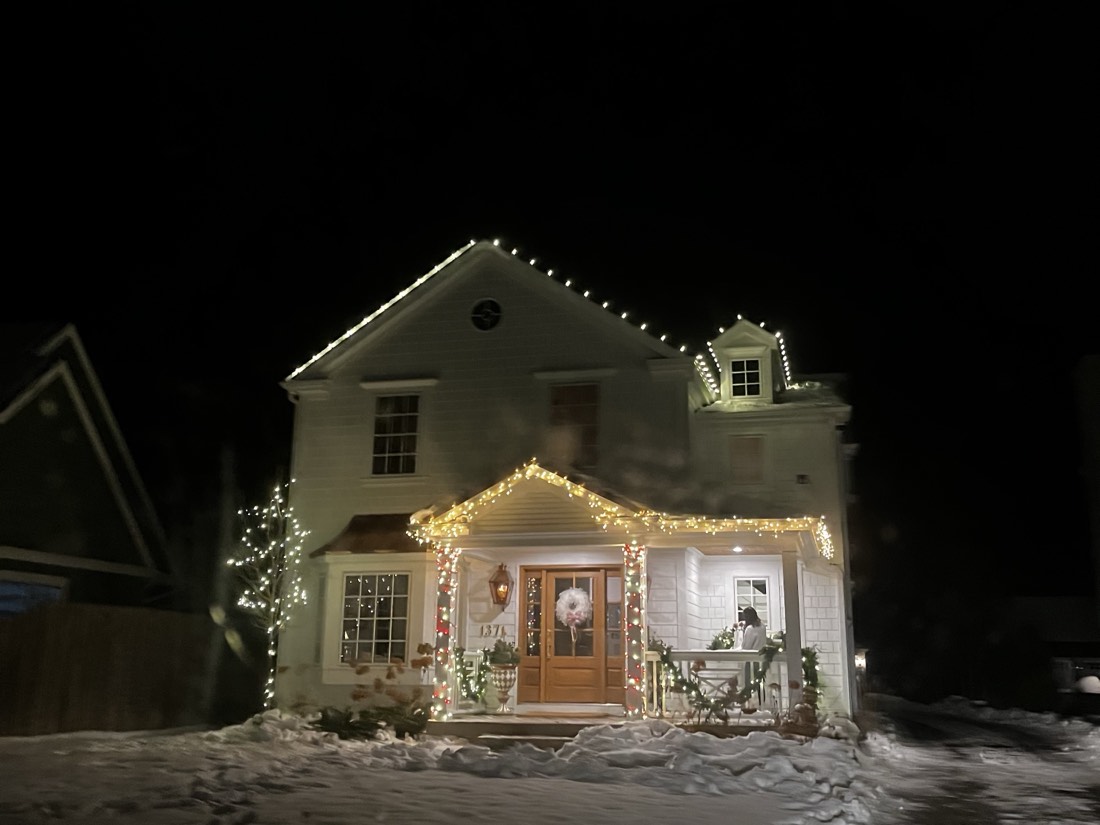 White farmhouse colonial after a snowfall, warm white C9 bulbs along the rooflines with garland and red accents across the front porch